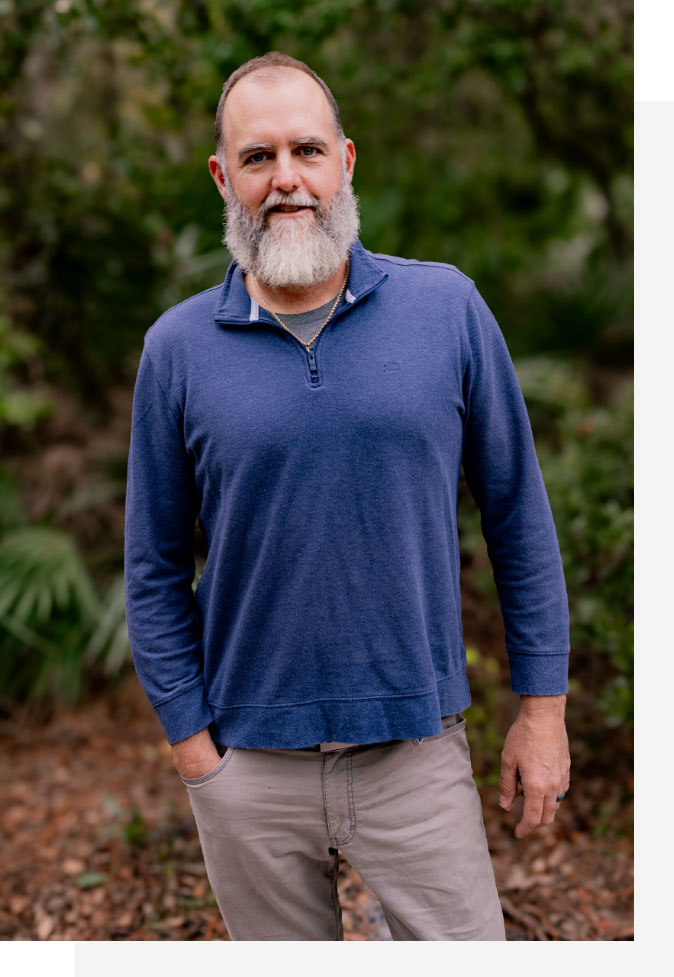 Photo of Jon Menke in blue long sleeve collared shirt with gray pants smiling with woods in background